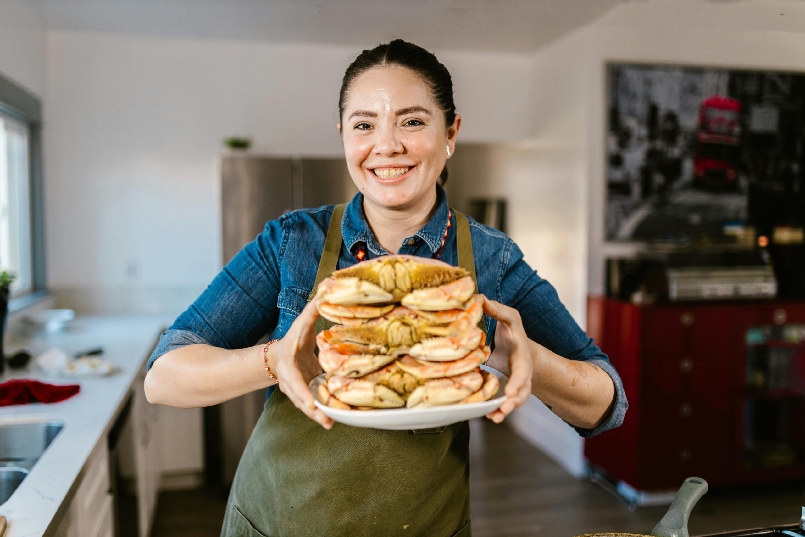 pexels-photo-10432741-10432741 Woman in a kitchen smiling, holding a stacked plate of crabs, portraying joy and culinary skills.